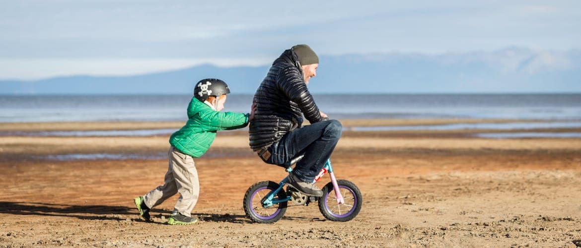 A child pushes an adult riding a tiny kids' bike along the Lake Tahoe shore